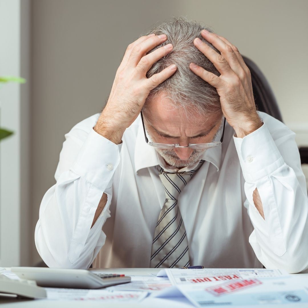 A stressed man holding his head in his hands, sitting at a desk with papers and a calculator